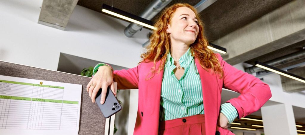 Vrouw in roze pak staat met mobiele telefoon in haar hand tegen een werkplek aan.