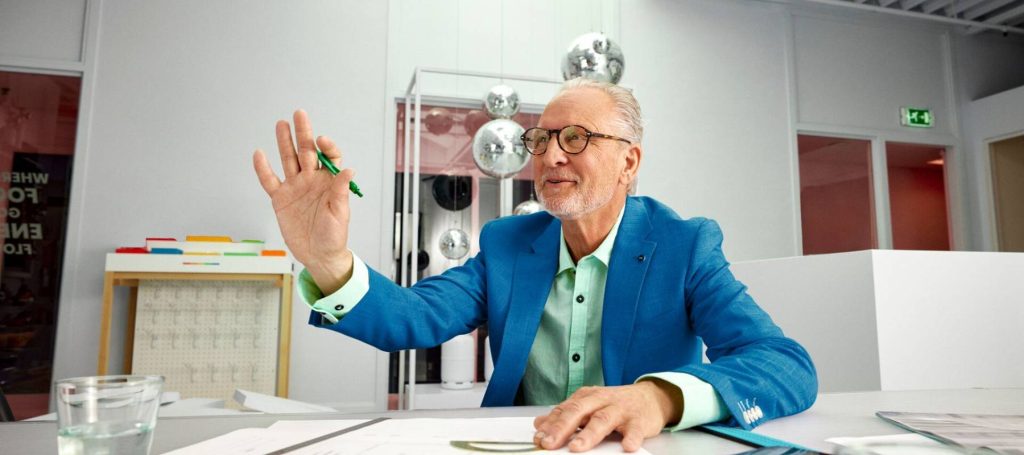 Man in blauw pak zit aan tafel en steekt zijn hand op